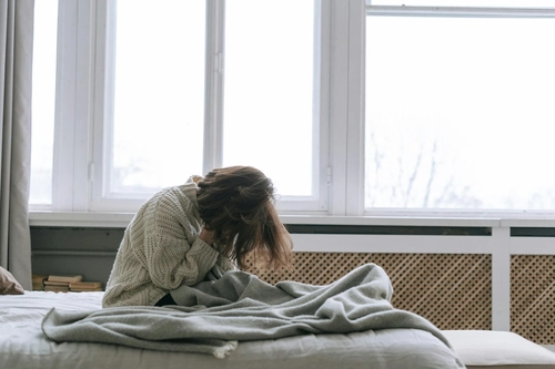 Depressed woman curling up with head bowed on bed beside big window
