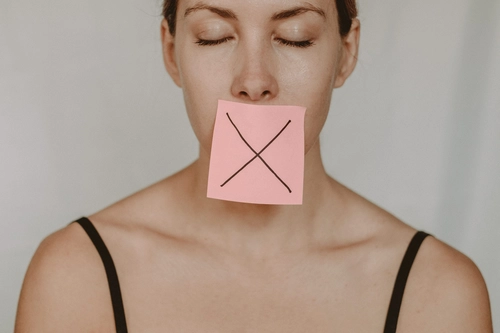 Woman with eyes closed, pink sticky note marked cross covering her mouth to symbolize silence