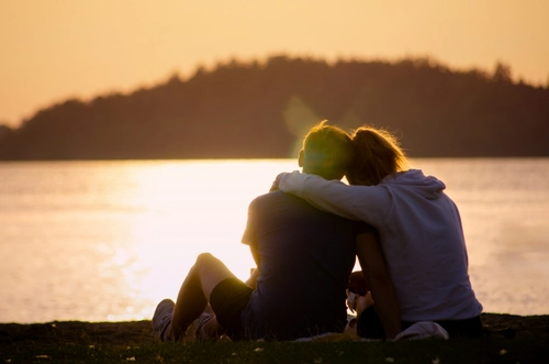 Couple sitting lakeside in sunset with one’s arm wrapped around the other