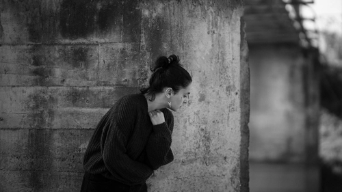 Anxious woman huddled against a rough concrete wall, shoulders hunched and body withdrawn.